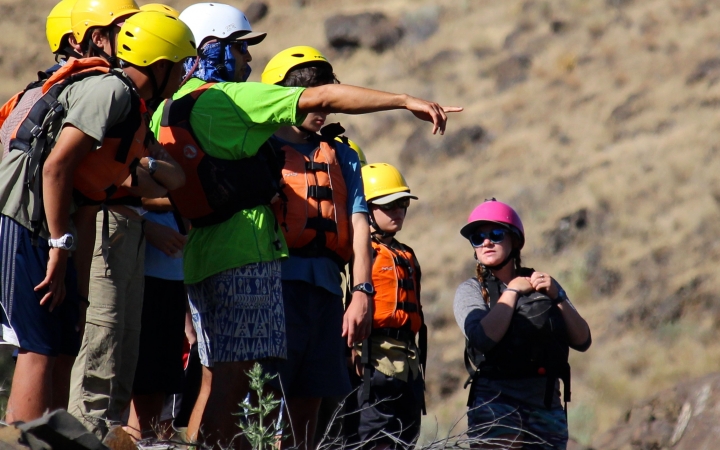 A group of students wearing life jackets and helmets stand in a line, possibly above a river. One of them is pointing at something in the distance.